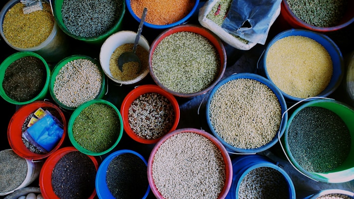 An assortment of various grains and seeds displayed in colorful plastic buckets. Each bucket is filled to the brim with different types of grains such as lentils, beans, and peas. A metal scoop is visible in one of the buckets, suggesting the grains are sold by weight. The vibrant colors of the buckets and the grains create an eye-catching display.