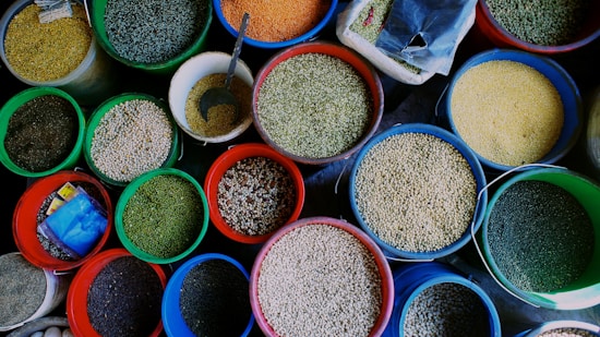 An assortment of various grains and seeds displayed in colorful plastic buckets. Each bucket is filled to the brim with different types of grains such as lentils, beans, and peas. A metal scoop is visible in one of the buckets, suggesting the grains are sold by weight. The vibrant colors of the buckets and the grains create an eye-catching display.