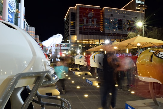 A night scene features a row of large football helmets on display in an outdoor area. The helmets represent various NFL teams, each placed on individual stands. People are walking around, creating a motion blur effect. Buildings with illuminated signs and advertisements are visible in the background, including a Hilton hotel. Bright lights and spotlights enhance the lively atmosphere.