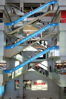Multiple levels of escalators crisscross inside a modern shopping mall. People are riding both up and down on the escalators, with some standing and others walking. Large blue advertisements for a phone brand are displayed along the escalators. Various store signs can be seen on different floors, providing a lively and bustling atmosphere.