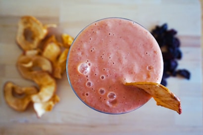 A glass filled with a thick, pink smoothie, topped with a slice of dried apple. To the left are several dried apple slices, and to the right, there seems to be a small pile of dark dried fruits, likely prunes or similar. The background is a light wooden surface, adding a natural and fresh vibe.