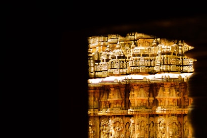 An ornate portion of an ancient temple featuring intricate carvings and golden hues is seen through a dark frame. The temple's facade is elaborately decorated with detailed sculptures and patterns, showcasing traditional architectural elements.