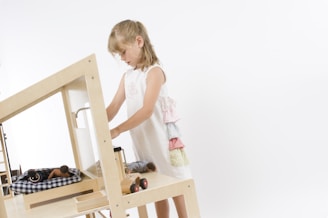 A baby girl in a floral dress playing with a wooden toy in a sunlit room.