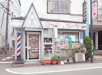 A quaint barbershop located on a street corner characterized by its retro aesthetic. The facade displays a vintage sign with Japanese lettering, featuring graphic scissors overhead. There's a classic barber pole on the wall, and the windows are adorned with posters and pictures. Potted plants line the sidewalk, enhancing the charming, old-fashioned atmosphere.