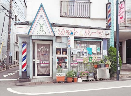 A quaint barbershop located on a street corner characterized by its retro aesthetic. The facade displays a vintage sign with Japanese lettering, featuring graphic scissors overhead. There's a classic barber pole on the wall, and the windows are adorned with posters and pictures. Potted plants line the sidewalk, enhancing the charming, old-fashioned atmosphere.