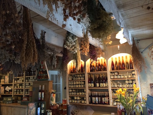 A rustic interior with shelves filled with various bottles and jars. Dried herbs and flowers hang from the ceiling, casting shadows and adding to the cozy, organic atmosphere. Warm lighting highlights the array of bottles, some filled with colorful liquids. A bouquet of fresh flowers sits prominently in the foreground.