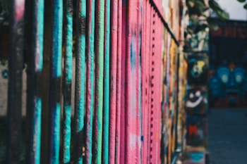 Brightly colored metal bars are covered with vibrant graffiti in an urban setting. A mix of teal, pink, and blue hues dominates the scene, highlighting the artistic expression of street art.