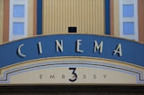 A vintage-style cinema sign with art deco elements. The blue and yellow arch displays the word 'CINEMA' prominently in bold letters above the smaller 'EMBASSY 3'. The design includes vertical ridges and geometric patterns along the edges.