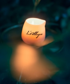 A glowing candle with the text 'L'Allegro' written on it, set against a blurred dark background that enhances the warm glow of the candlelight.