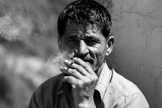 A black and white portrait of a man smoking, with smoke curling around his face. His expression seems contemplative, and he is wearing a collared shirt. The background is blurred and rustic.