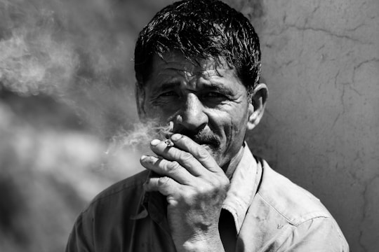 A black and white portrait of a man smoking, with smoke curling around his face. His expression seems contemplative, and he is wearing a collared shirt. The background is blurred and rustic.