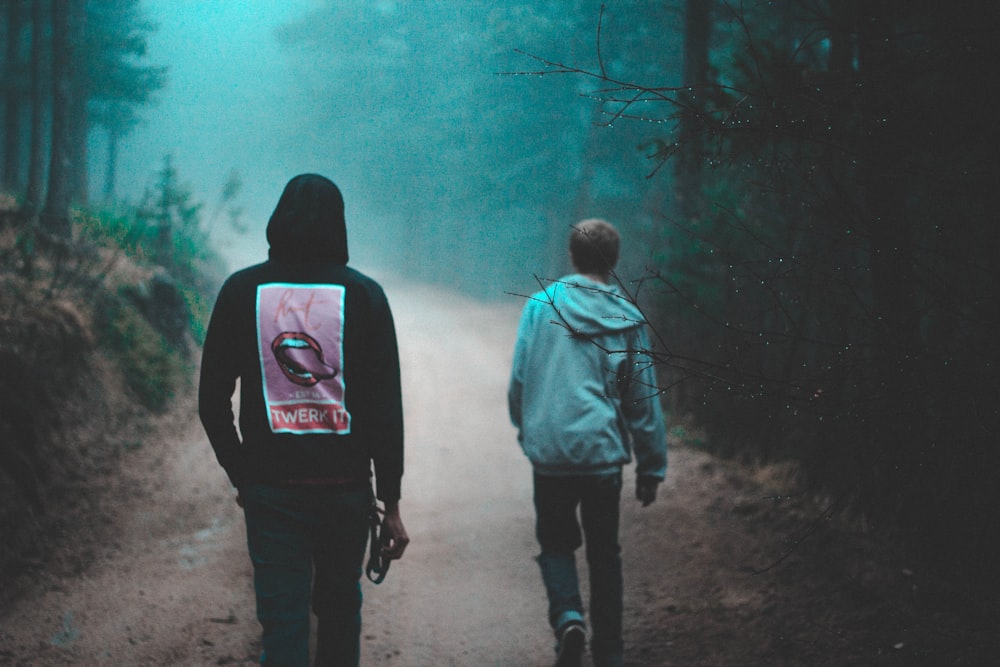 two person walking on dirt road between green trees