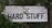 A rustic wooden sign with the text 'Industrial Furniture' and 'Hard Stuff' written in bold, black letters. The background consists of weathered white wood planks. The sign is partially covered with overgrown green foliage and leaves, giving it a natural, earthy appearance.