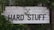 A rustic wooden sign with the text 'Industrial Furniture' and 'Hard Stuff' written in bold, black letters. The background consists of weathered white wood planks. The sign is partially covered with overgrown green foliage and leaves, giving it a natural, earthy appearance.
