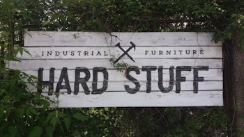 A rustic wooden sign with the text 'Industrial Furniture' and 'Hard Stuff' written in bold, black letters. The background consists of weathered white wood planks. The sign is partially covered with overgrown green foliage and leaves, giving it a natural, earthy appearance.