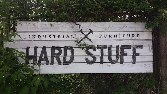 A rustic wooden sign with the text 'Industrial Furniture' and 'Hard Stuff' written in bold, black letters. The background consists of weathered white wood planks. The sign is partially covered with overgrown green foliage and leaves, giving it a natural, earthy appearance.
