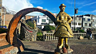 A golden statue of a person dressed in traditional attire stands prominently in an outdoor plaza. The statue is positioned under an arched stone structure surrounded by cobblestone pathways. In the background, there are buildings with rectangular windows and a well-maintained garden area featuring another smaller statue. The scene is vibrant with blue skies overhead and a few clouds.