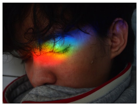 A person with closed eyes is seen with a vibrant rainbow of colors projected onto their face, positioned across the eyelid. The colors range from red, orange, yellow, green, blue, to purple. A tear appears to trail down the cheek, adding a sense of emotion to the scene.