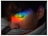 A person with closed eyes is seen with a vibrant rainbow of colors projected onto their face, positioned across the eyelid. The colors range from red, orange, yellow, green, blue, to purple. A tear appears to trail down the cheek, adding a sense of emotion to the scene.