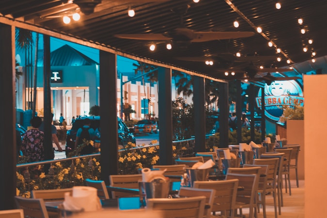 An outdoor dining area with wooden tables and chairs is set under string lights, creating a warm and inviting ambiance. Adjacent to the dining area, a street scene with multiple people walking by and a view of a building with modern architecture can be seen. The logo of a restaurant is visible in the background, indicating a lively, urban setting.