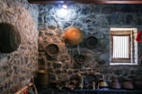 A rustic room with stone walls adorned with a variety of traditional brass and copper utensils. On the wall, baskets and trays are hung for decoration. A small window lets in natural light, illuminating a collection of pots and kitchen implements on a wooden shelf below.
