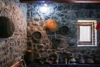 A rustic room with stone walls adorned with a variety of traditional brass and copper utensils. On the wall, baskets and trays are hung for decoration. A small window lets in natural light, illuminating a collection of pots and kitchen implements on a wooden shelf below.