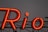 Large, bright red neon sign against a dark background, forming the word 'Rio' with distinct glowing letters, mounted on a metal structure.