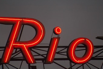 Large, bright red neon sign against a dark background, forming the word 'Rio' with distinct glowing letters, mounted on a metal structure.
