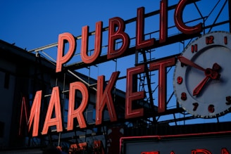 A neon sign displaying the words 'Public Market' with a large clock showing the time. The red letters and clock stand out against a backdrop that suggests an outdoor market setting. The sky in the background is a deep blue.