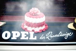 A small, knitted or crocheted item resembling a cake is placed on a reflective surface. Raindrops are visible, giving a wet and slightly blurred look. The text 'OPEL der Zuverlässige' is displayed prominently, blending with the background.