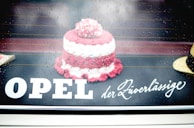 A small, knitted or crocheted item resembling a cake is placed on a reflective surface. Raindrops are visible, giving a wet and slightly blurred look. The text 'OPEL der Zuverlässige' is displayed prominently, blending with the background.