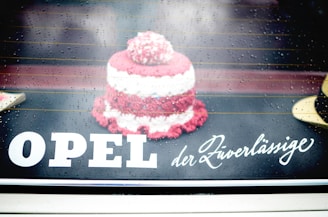 A small, knitted or crocheted item resembling a cake is placed on a reflective surface. Raindrops are visible, giving a wet and slightly blurred look. The text 'OPEL der Zuverlässige' is displayed prominently, blending with the background.