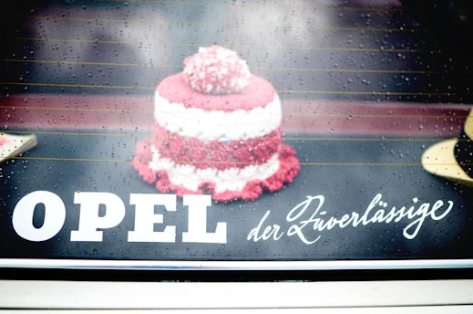 A small, knitted or crocheted item resembling a cake is placed on a reflective surface. Raindrops are visible, giving a wet and slightly blurred look. The text 'OPEL der Zuverlässige' is displayed prominently, blending with the background.