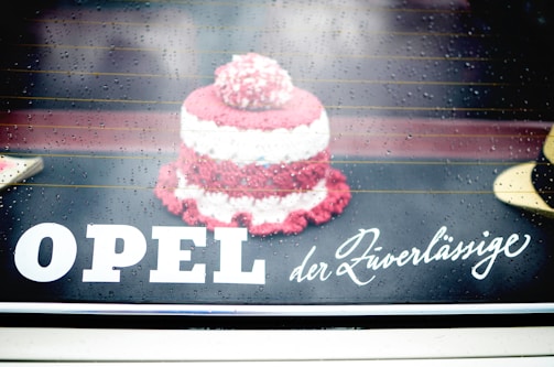 A small, knitted or crocheted item resembling a cake is placed on a reflective surface. Raindrops are visible, giving a wet and slightly blurred look. The text 'OPEL der Zuverlässige' is displayed prominently, blending with the background.