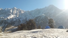 A cozy Himalayan tent pitched against a backdrop of snow-capped Kedarnath peaks under a clear blue sky.