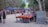 A vibrant outdoor market in a wooded area with various vendors displaying their goods on tables. Colorful textiles, handmade crafts, and souvenirs are visible. Several people are browsing and walking along the path, which is lined with purple umbrellas providing shade.