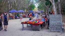 A vibrant outdoor market in a wooded area with various vendors displaying their goods on tables. Colorful textiles, handmade crafts, and souvenirs are visible. Several people are browsing and walking along the path, which is lined with purple umbrellas providing shade.
