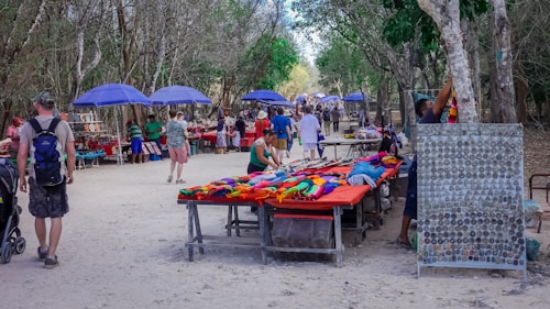 A vibrant outdoor market in a wooded area with various vendors displaying their goods on tables. Colorful textiles, handmade crafts, and souvenirs are visible. Several people are browsing and walking along the path, which is lined with purple umbrellas providing shade.