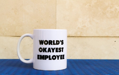 A white mug with the words 'WORLD'S OKAYEST EMPLOYEE' printed in bold black letters. The mug is placed on a blue textured surface with a beige stone wall in the background.