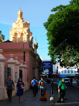 A vibrant street scene features a group of people walking near a pink colonial-style building with ornate architectural details. A street cleaner in a green uniform is sweeping the road, and a large green tree provides shade. In the background, a blue bus and a commercial building can be seen, along with a clear blue sky.