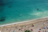 A sandy beach with many people sunbathing and enjoying leisure activities. The water is a clear turquoise blue, and a few people are swimming. A small boat is visible on the water near the shore. Colorful umbrellas and towels dot the beach.