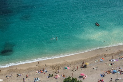 A sandy beach with many people sunbathing and enjoying leisure activities. The water is a clear turquoise blue, and a few people are swimming. A small boat is visible on the water near the shore. Colorful umbrellas and towels dot the beach.