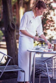 A person with blonde hair, dressed in a white shirt and apron, is setting or clearing a table outdoors. A metal table and chair are present, surrounded by greenery. The person is handling a small item, possibly cutlery.