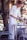 A person with blonde hair, dressed in a white shirt and apron, is setting or clearing a table outdoors. A metal table and chair are present, surrounded by greenery. The person is handling a small item, possibly cutlery.