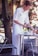 A person with blonde hair, dressed in a white shirt and apron, is setting or clearing a table outdoors. A metal table and chair are present, surrounded by greenery. The person is handling a small item, possibly cutlery.