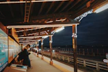 A dimly lit train platform with a wooden overhang, illuminated by string lights, creating a warm ambient glow. A person in a dark coat and scarf stands on the left side, seemingly focused on a phone. The platform looks out over a roadway next to a large body of water under a clouded night sky. Posters and signs line the walls next to the person.