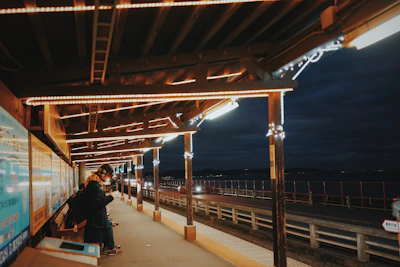A dimly lit train platform with a wooden overhang, illuminated by string lights, creating a warm ambient glow. A person in a dark coat and scarf stands on the left side, seemingly focused on a phone. The platform looks out over a roadway next to a large body of water under a clouded night sky. Posters and signs line the walls next to the person.