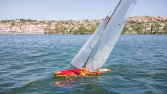 A model sailboat with a red hull is navigating on a body of water. The sails are taut, indicating a gentle breeze. In the background, a shoreline is visible with residential buildings and greenery.