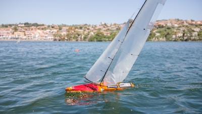 A model sailboat with a red hull is navigating on a body of water. The sails are taut, indicating a gentle breeze. In the background, a shoreline is visible with residential buildings and greenery.