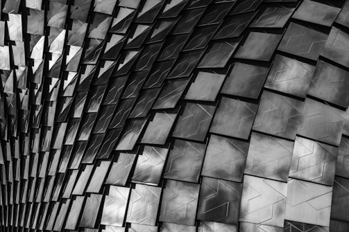 A close-up view of a textured surface composed of overlapping, metallic tiles arranged in a wave-like pattern. The tiles have a geometric design with a mix of dark and light shades, creating a sense of depth and movement.